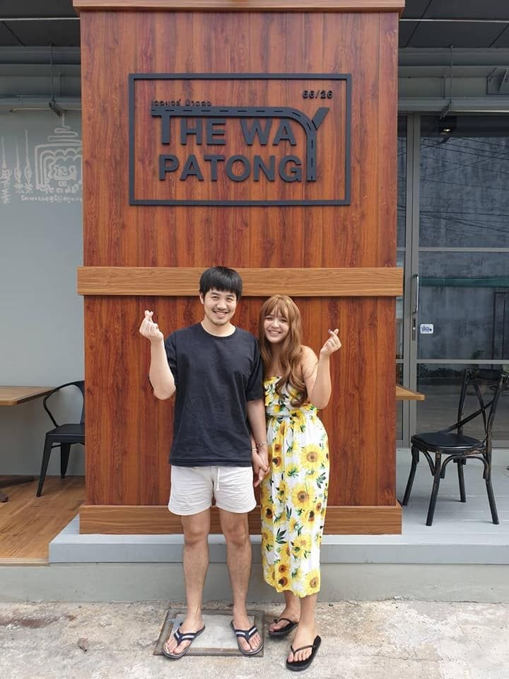 A couple making heart signs in front of The Way Patong wooden logo sign
